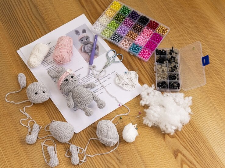 Anleitung, Wollknäuel in Rosa und Weiß, halbfertige gehäkelte Stofftierteile und Perlen auf Holzuntergrund ausgebreitet. | © BILD der FRAU/Photoproduction Bernd Hanselmann
