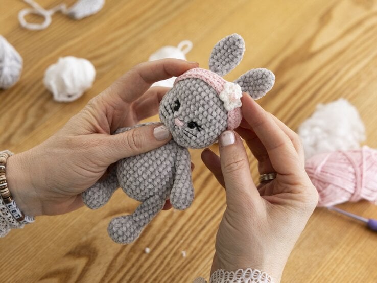 Hände präsentieren ein graues gehäkeltes Stofftier mit rosa Stirnband und Blume auf hellem Holztisch. | © BILD der FRAU/Photoproduction Bernd Hanselmann