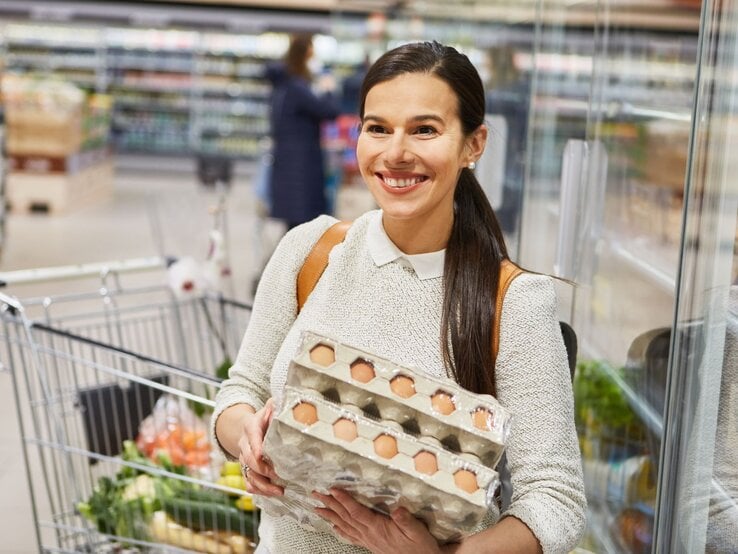 Eine junge Frau mit braunem Haar und weißem Pullover hält lächelnd zwei Eierkartons in einem Discounter.