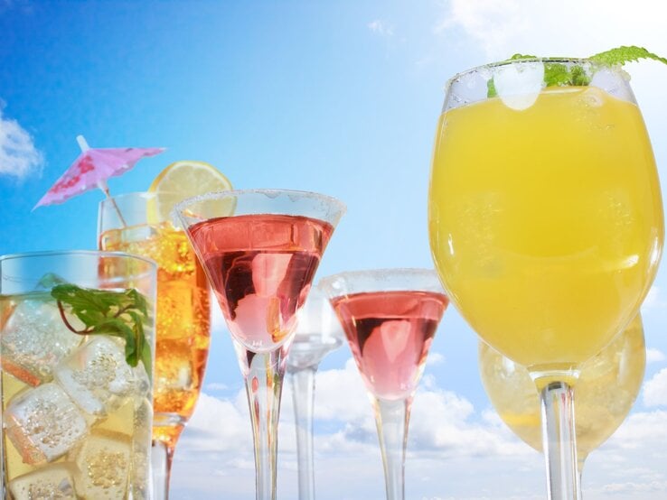 Bunte Cocktails mit Früchten, Minze und Eiswürfeln stehen vor strahlend blauem Himmel und weißen Wolken. | © Getty Images/Melpomenem