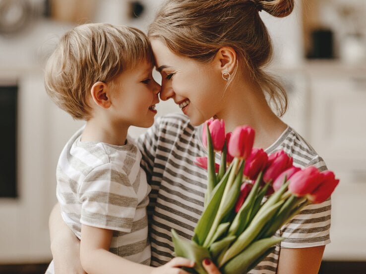 Eine Mutter nimmt ihren kleinen Sohn lächelnd in den Arm der ihr einen Strauß roter Tulpen schenkt. Man sieht beide von der Seite, wie sie sich anlächeln.