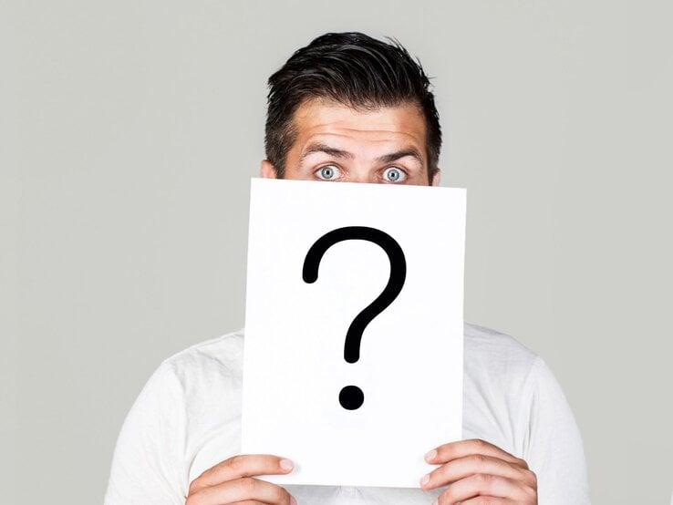 Ein junger Mann mit stechend blauen Augen hält ein Schild mit großem Fragezeichen vor sein Gesicht auf grauem Hintergrund.
