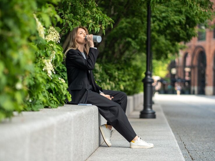 Eine Frau in schwarzem Hosenanzug und weißen Sneakern sitzt entspannt auf einer Steinmauer und trinkt aus einem Becher vor grünem Gebüsch.