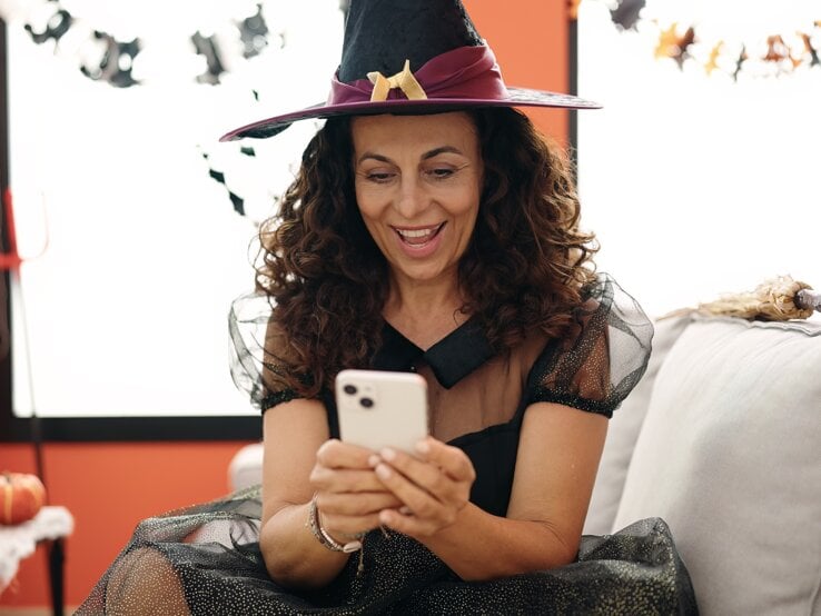 Ein festlich dekorierter Raum mit Kürbissen, Kerzen und Spinnweben, dazu eine Person mit Hexenkostüm und Smartphone in der Hand.