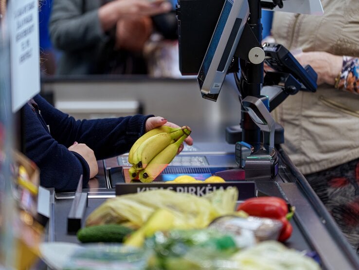 Gelbe Bananen werden an einer belebten Supermarktkasse gescannt, vorne liegen Gurke und Paprika, hinten Kleidung und Hände sichtbar.