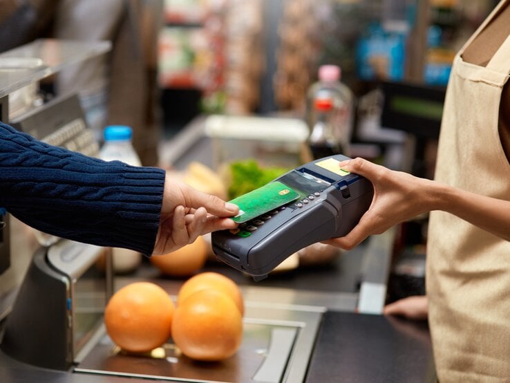 Eine Person in blauem Pullover hält eine grüne Kreditkarte über ein Kartenterminal, im Hintergrund sind frische Lebensmittel zu sehen.