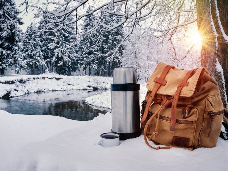 Rucksack und Thermoskanne stehen im Schnee vor winterlichem Wald und Fluss, Sonnenlicht fällt durch verschneite Äste.
