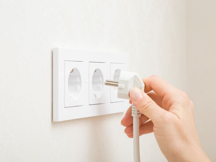 Eine Hand steckt einen weißen Stecker in eine von drei weißen Steckdosen an einer hellen Wand.