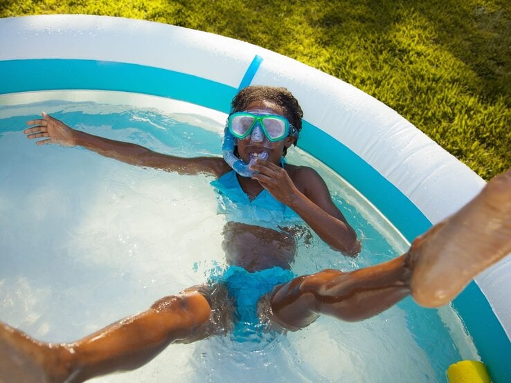 Ein fröhliches Mädchen in blauer Badebekleidung und Taucherbrille planscht mit ausgestreckten Armen im aufblasbaren Pool im Garten.