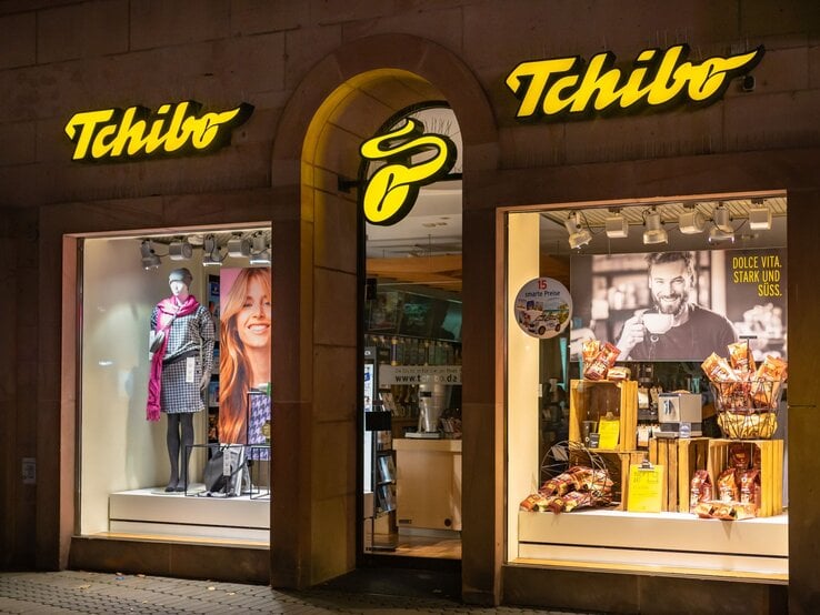 Tchibo-Schaufenster bei Nacht mit Modepuppe in karierter Jacke, rotem Schal und Werbung für Kaffee mit lächelndem Mann im Hintergrund.
