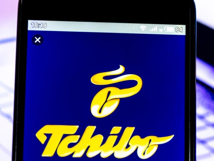 Tchibo-Logo auf einem Smartphone-Bildschirm vor einem unscharfen Laptop, der einen Kursverlauf anzeigt.