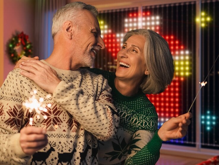 Ein älteres Paar in Weihnachtspullovern lacht sich liebevoll an und hält Wunderkerzen, im Hintergrund festliche Lichterdeko.
