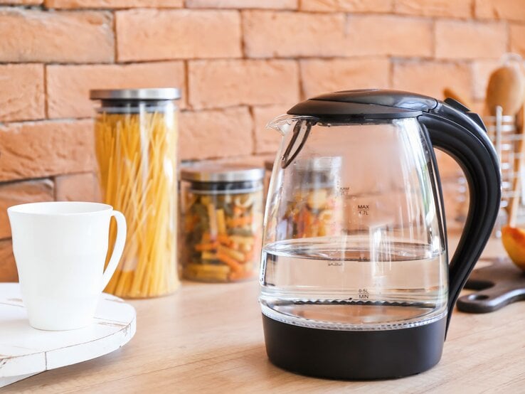 Ein transparenter Wasserkocher mit schwarzem Griff steht auf einer Holzarbeitsplatte, daneben eine weiße Tasse und Gläser mit Nudeln vor einer Ziegelwand.
