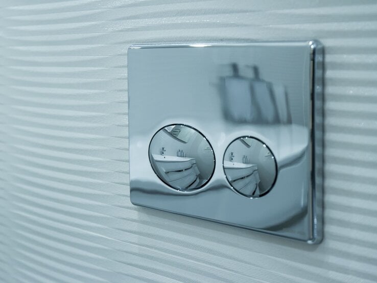 Verchromtes WC-Spülsystem mit zwei runden Tasten spiegelt ein modernes Badezimmer, montiert auf geriffelter weißer Wand.