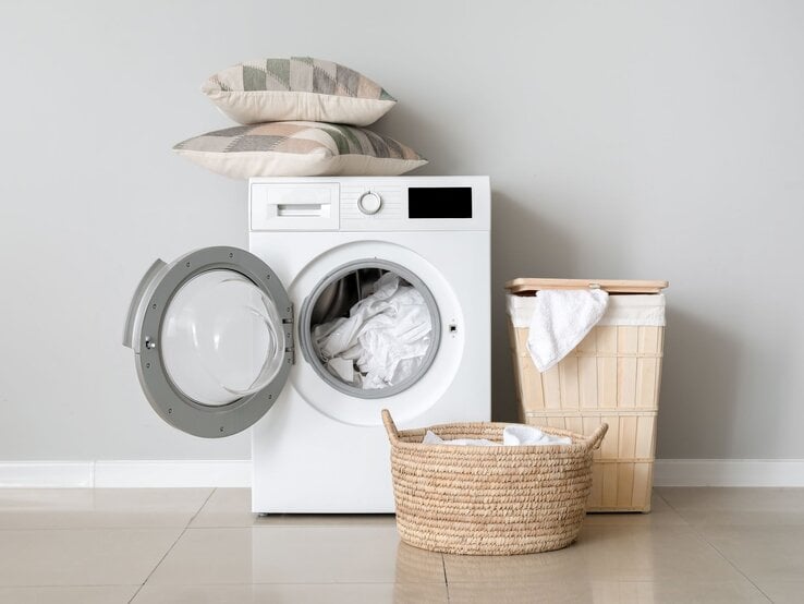 Eine weiße Waschmaschine mit offener Tür, aus der weiße Wäsche ragt, daneben zwei Wäschekörbe aus Korbgeflecht vor grauer Wand.