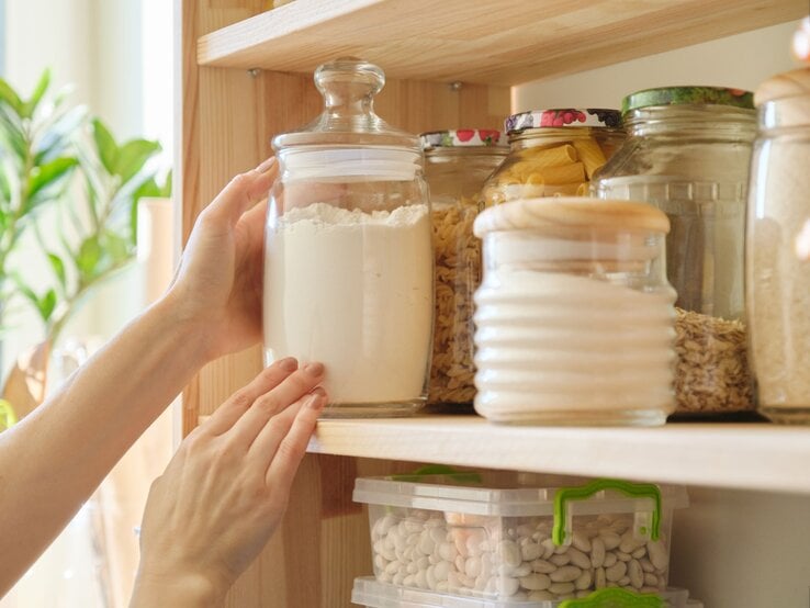 Eine Hand greift in einer hellen Küche nach einem großen Glas mit Mehl, das in einem Holzregal zwischen weiteren Vorratsgläsern steht.