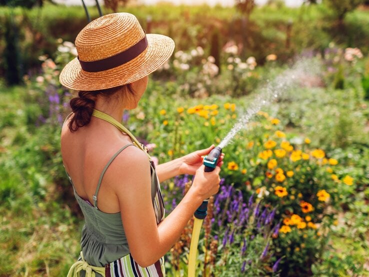 Junge Frau mit Strohhut gießt an einem sonnigen Tag bunte Sommerblumen im üppigen Garten mit einem Wasserschlauch.