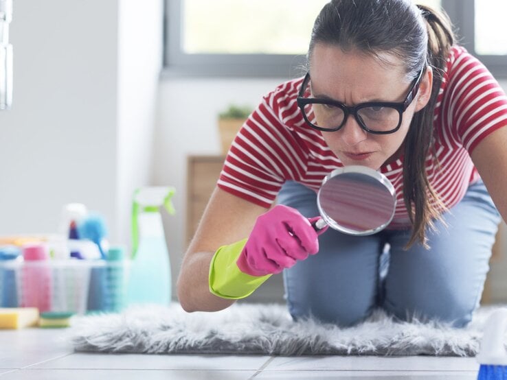 Junge Frau mit Brille und Putzhandschuhen kniet ernsthaft auf dem Boden und sucht mit Lupe nach Schmutz im Badezimmer.
