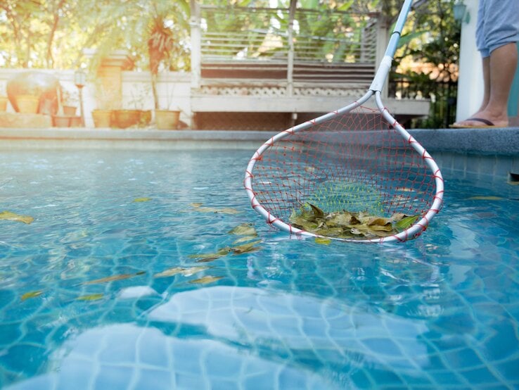Ein Mensch in Flipflops fischt mit einem Kescher herabgefallene Blätter aus einem blauen Pool bei sanftem Sonnenlicht.