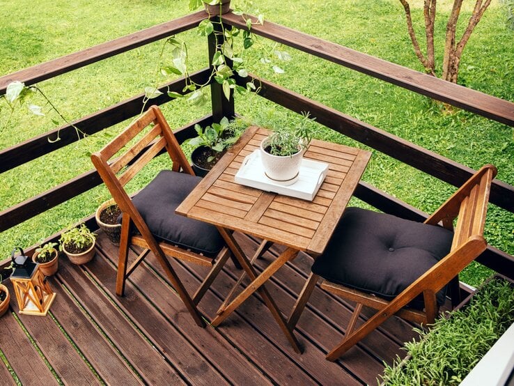 Zwei Holzklappstühle mit schwarzen Polstern stehen an einem kleinen Holztisch auf einer Terrasse mit Kräutertöpfen und Grünpflanzen.