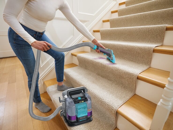 Junge Frau in weißem Langarmshirt und Jeans reinigt konzentriert mit Teppichreiniger einen lilafarbenen Fleck auf beiger Treppe. | © SharkNinja