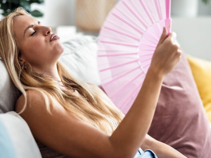 Junge blonde Frau mit müdem Gesichtsausdruck fächelt sich auf dem Sofa mit rosa Fächer Luft zu, sichtbar erschöpft von Hitze.