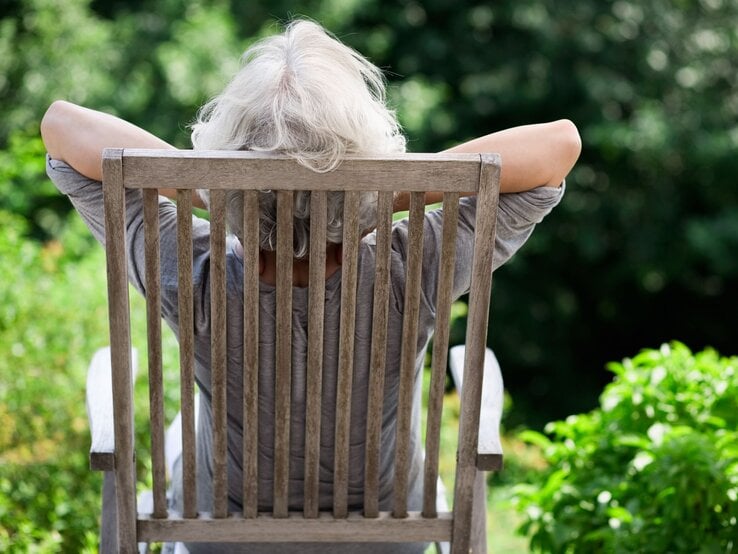 Eine grauhaarige ältere Person lehnt sich entspannt im Holzstuhl zurück und blickt auf üppiges, grünes Gartenlaub.