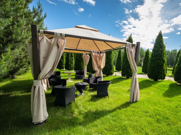 Ein luftiger Gartenpavillon mit offenen Vorhängen lädt inmitten symmetrisch gepflanzter Koniferen zum Verweilen ein.