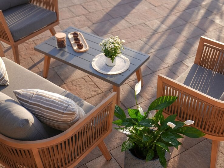 Elegante Gartenlounge mit Holzmöbeln und grauen Polstern um einen Tisch mit Muffins, Blumen und Keksdose in der Abendsonne.