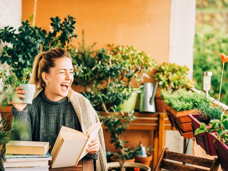Junge blonde Frau in Strickpullover lacht mit Buch und Tasse auf grün bepflanztem Balkon voller Kräuter und Blumentöpfe.
