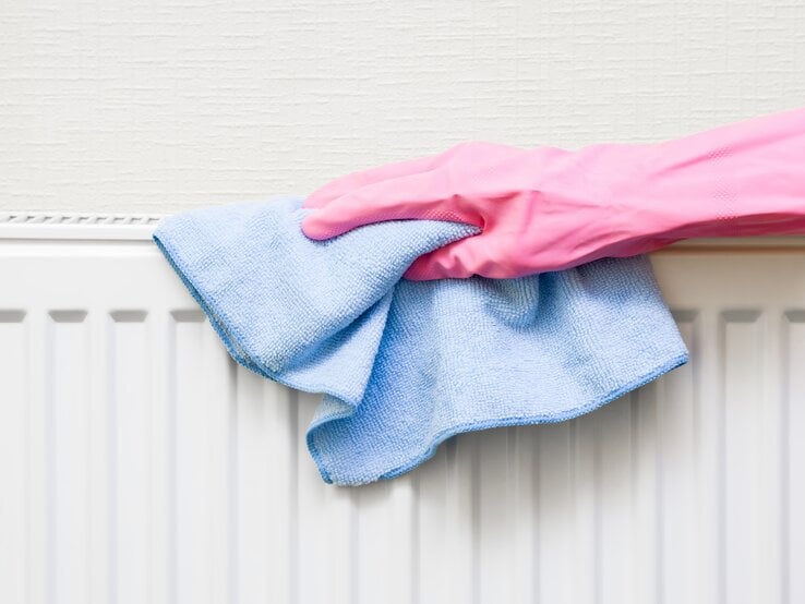 Eine Hand in pinkem Gummihandschuh wischt mit einem blauen Mikrofasertuch einen weißen Heizkörper vor cremefarbener Wand ab.