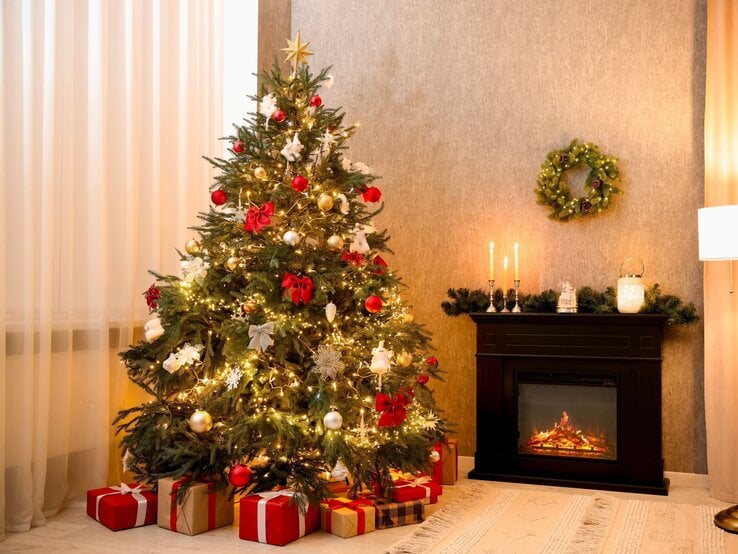 Geschmückter Weihnachtsbaum mit roten Schleifen und glänzenden Kugeln, daneben leuchtender Kamin mit Kerzen und Tannendekor.