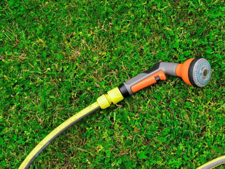 Gartenschlauch auf grünem Rasen mit orangem Sprühkopf und zwei gelben Schwämmen im blauen Kreis daneben.