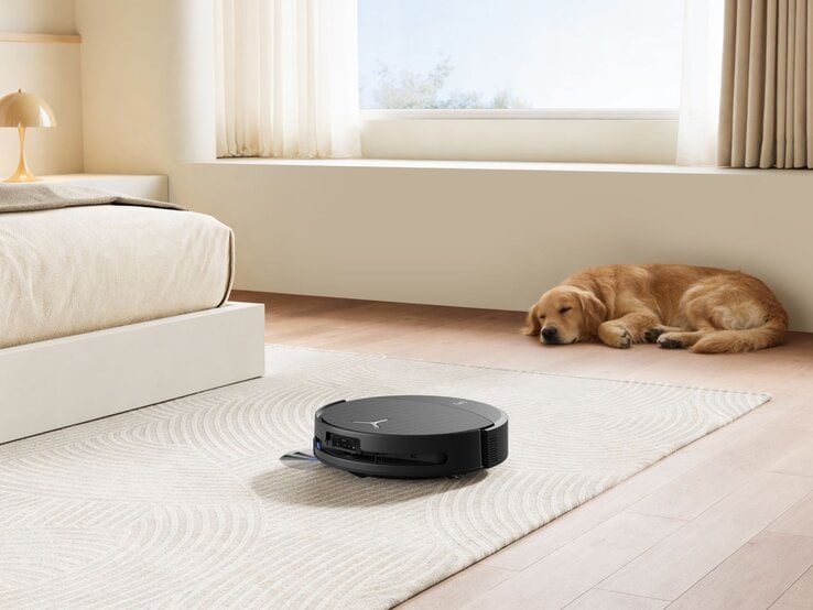 Ein Hund schläft friedlich auf einem Holzboden neben einem Teppich, während ein schwarzer Staubsaugerroboter den Raum reinigt. | © ECOVACS
