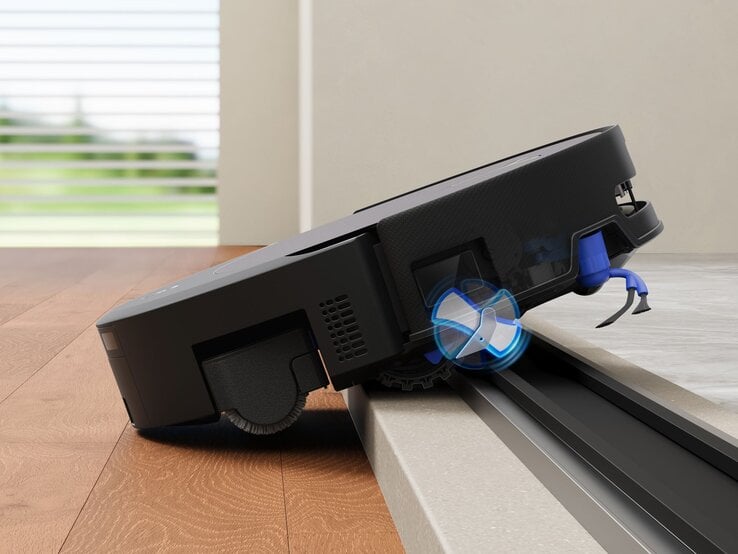 Schwarzer Staubsaugerroboter überwindet geschickt eine Schwelle zwischen Holz- und Fliesenboden, blaue Bürsten in Aktion. | © ECOVACS