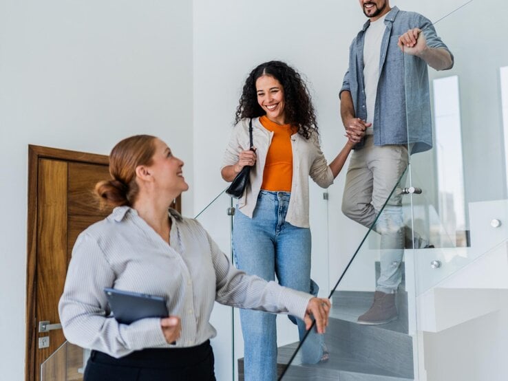 Eine Maklerin zeigt einer Frau und einem Mann eine Wohnung. Alle gehen hintereinander die Treppe runter. 