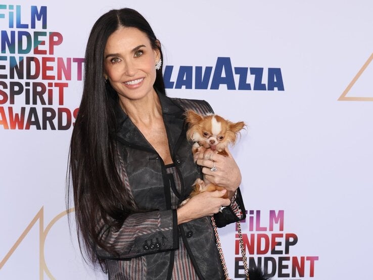 Dunkelhaarige, lächelnde Frau in gemusterter Jacke hält kleinen braun-weißen Hund auf rotem Teppich mit bunter Award-Wand.