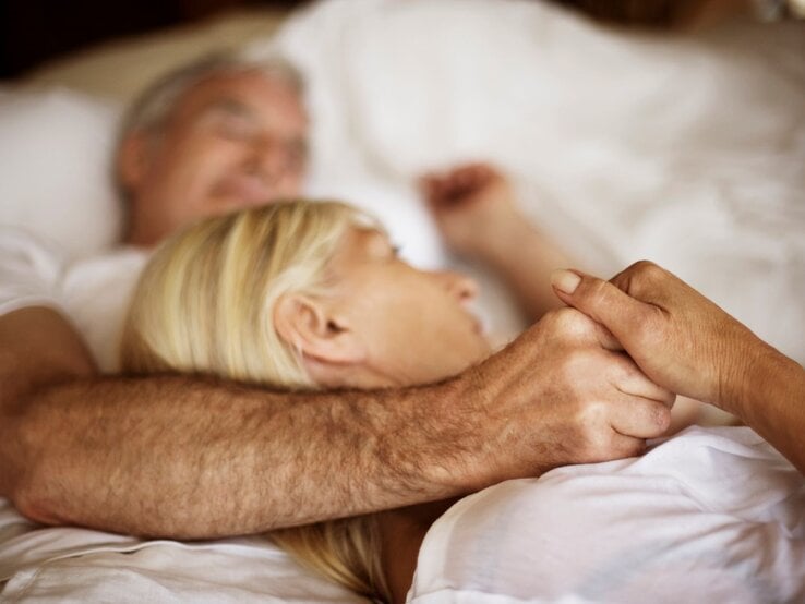 Ein älteres Paar liegt eng umschlungen in einem weißen Bett, hält sich liebevoll an den Händen und wirkt entspannt und geborgen.