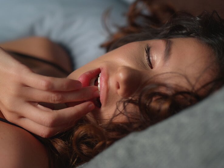 Eine junge Frau mit dunklen Locken liegt mit geschlossenen Augen auf einem Kissen und berührt sanft ihre Lippen mit den Fingern.