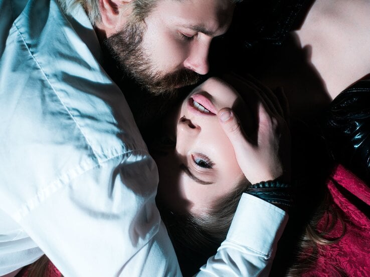 Ein bärtiger Mann in weißem Hemd hält das Gesicht einer jungen Frau mit offenem Blick auf dunklem Untergrund fest.