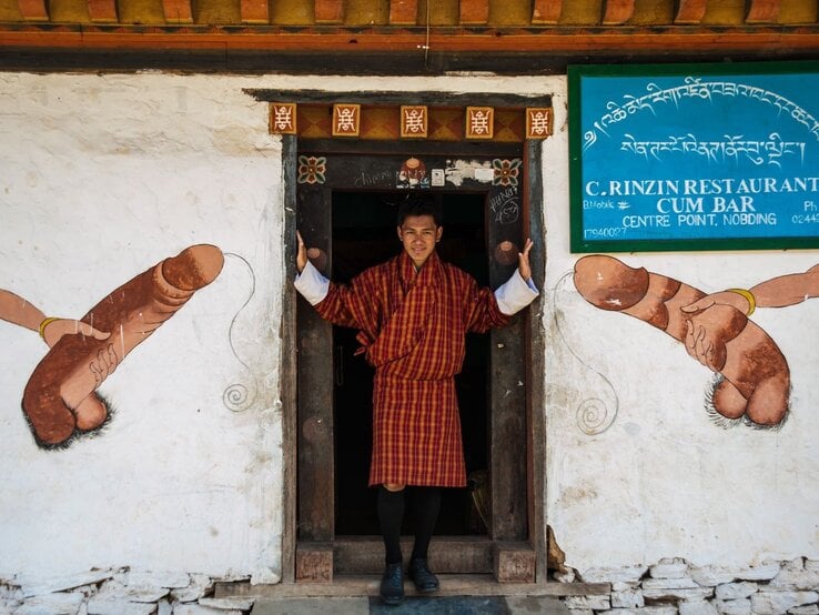 Junger Bhutaner in rot-gelbem Gho steht lächelnd im Türrahmen, flankiert von traditionellen Phallusmalereien an weißer Wand.