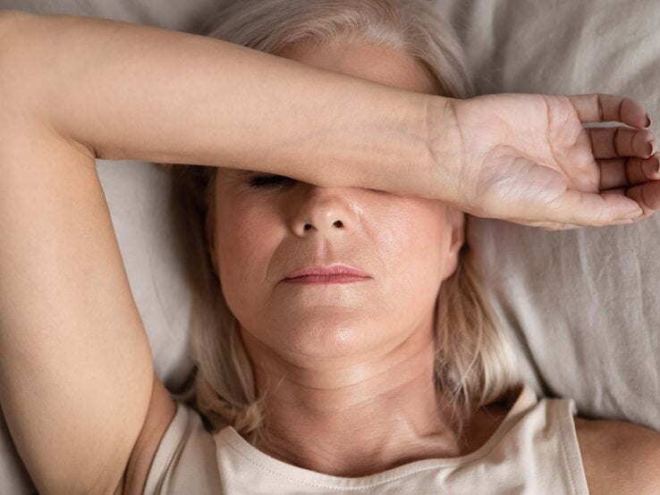 Ältere Frau mit blondem Haar liegt mit ernster Miene im Bett, trägt ein beiges Top und bedeckt ihre Augen mit dem rechten Unterarm.