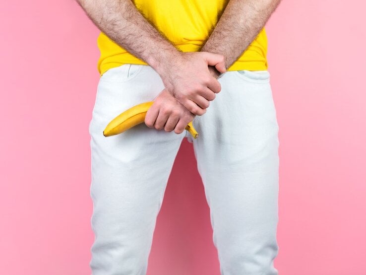 Junger Mann in gelbem T-Shirt hält eine Banane provokant vor seinem Schritt, vor rosa Hintergrund und weißer Hose.