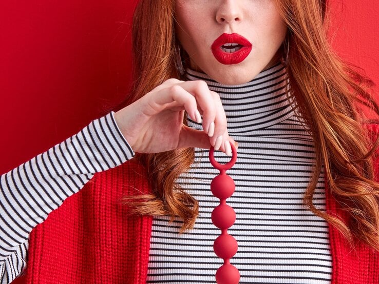 Junge Frau mit roten Lippen und langen roten Haaren hält rote Liebeskugeln vor rot gestreiftem Hintergrund in Brusthöhe.