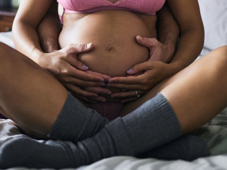 Junge schwangere Frau in rosa Unterwäsche sitzt auf Bett, Partner umarmt sie liebevoll, beide Hände auf Babybauch.