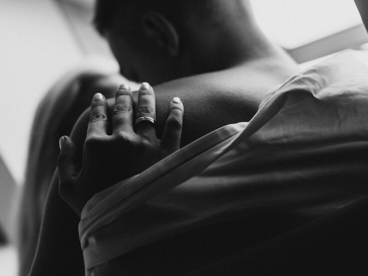 Nahaufnahme eines jungen Paares in inniger Umarmung, eine Hand mit Ring ruht zärtlich auf der nackten Schulter.