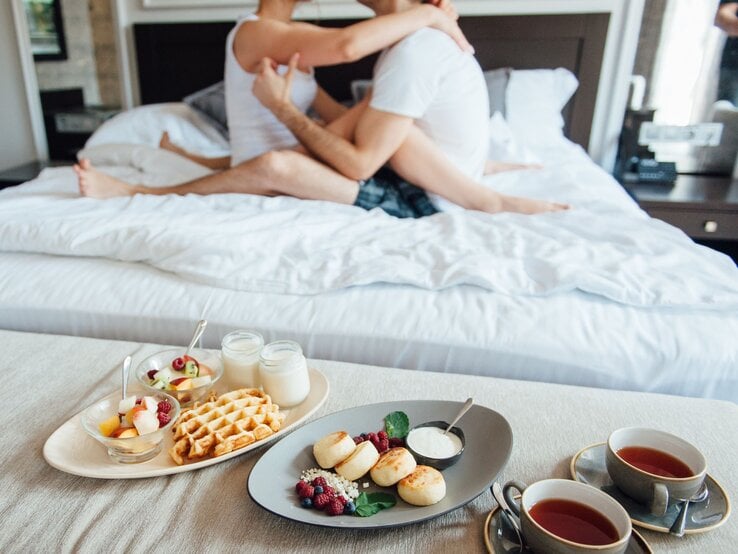 Zwei junge Männer in weißen Shirts umarmen sich innig auf einem Bett, davor ein Frühstückstablett mit Tee, Obst und Waffeln.