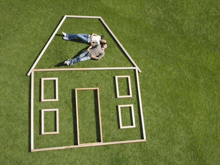 Junges Paar liegt träumend im Obergeschoss eines aus Holzrahmen geformten Hauses auf einer sattgrünen Wiese.