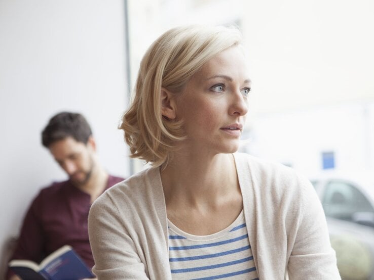 Frau schaut gedankenverloren aus dem Fenster. Im Hintergrund sitzt ein Mann und liest ein Buch, ohne ihre Anwesenheit zu bemerken.
