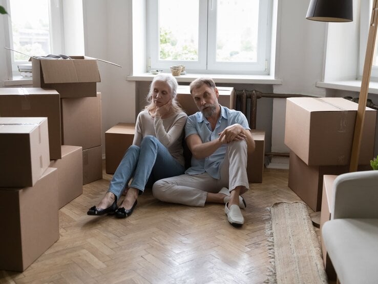 Paar sitzt erschöpft zwischen zahlreichen Umzugskartons in einem Raum. Die beiden Personen, eine Frau und ein Mann, scheinen eine Pause zu machen und wirken nachdenklich.  | © Adobe Stock / fizkes